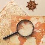 Top view of round magnifying glass on paper maps placed on beige background with wooden wind rose in light room